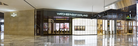 Luxury watch store with glass storefront and bright signage, displaying Rolex and Patek Philippe brands.