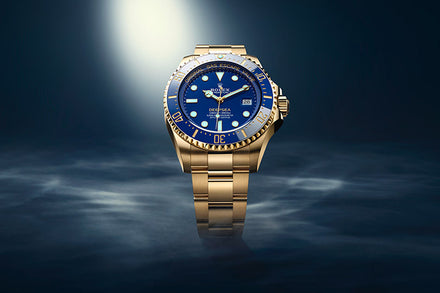 A gold Rolex Deepsea watch with a blue face and bezel is spotlighted against a dark, misty background.