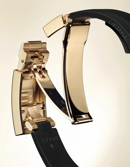 Close-up of a gold Rolex watch clasp with a black Oysterflex rubber strap on a light background.