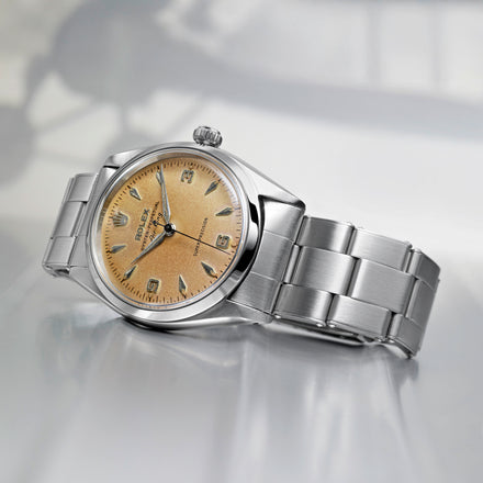 A silver Rolex wristwatch with a gold textured dial displayed on a reflective surface.