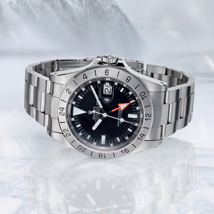 A stainless steel Rolex watch with a black dial and orange hand, displayed on an icy surface.