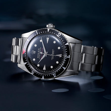 A stainless steel Rolex wristwatch with a black dial and bezel, displayed on a dark reflective surface.