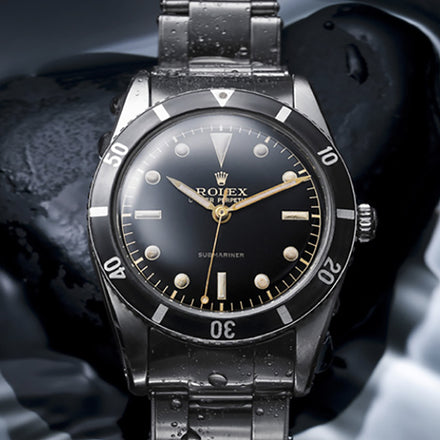 A vintage Rolex Submariner watch with a black dial and metal strap, shown close-up with water droplets.
