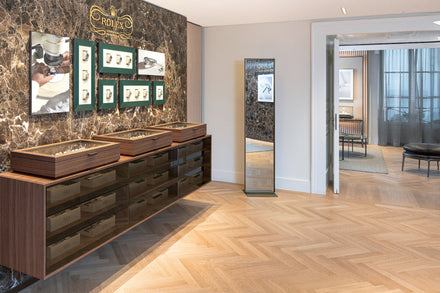 A luxury watch store with display cases, framed watch images, marble wall, and a full-length mirror.