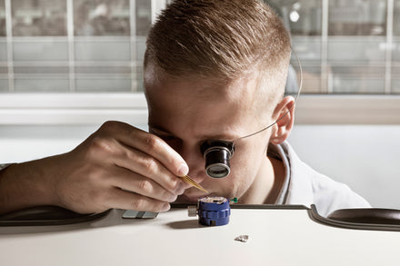Person using a magnifying loupe and tool to work on a small watch mechanism at a table.