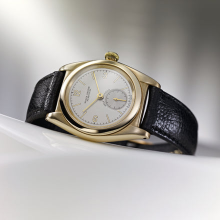 A vintage gold wristwatch with a black leather strap and silver dial, displayed on a white curved surface.