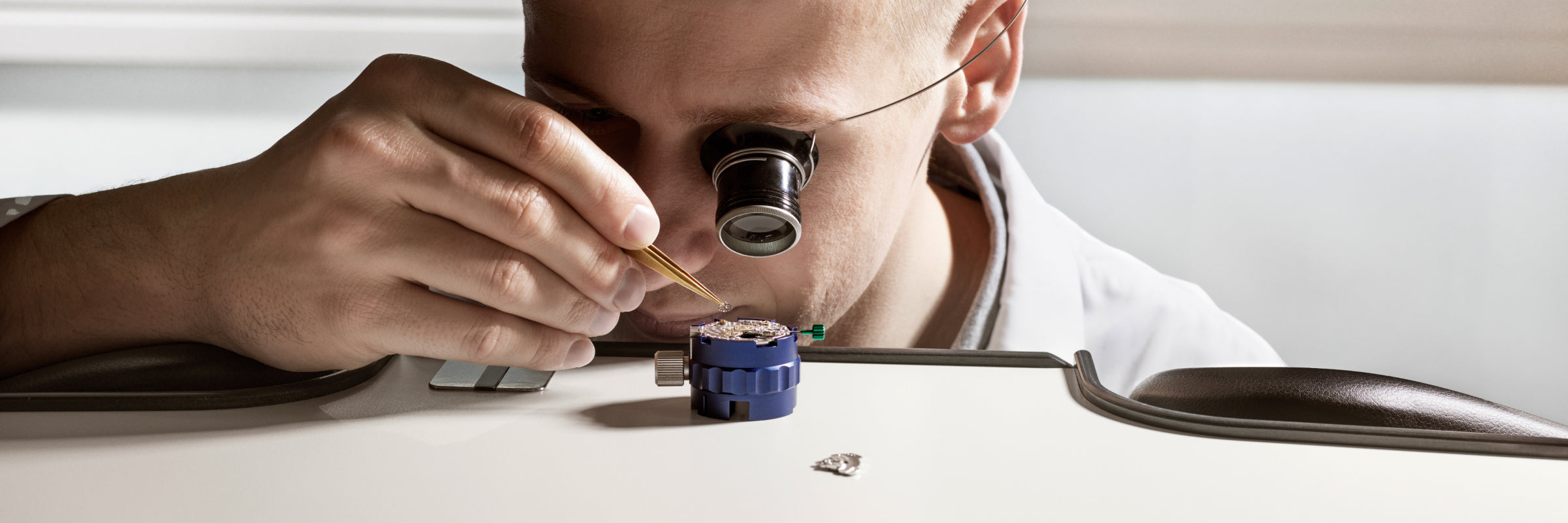 A watchmaker uses tweezers and a loupe to work on a small watch mechanism at a table.
