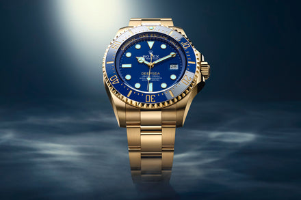 Gold Rolex Deepsea watch with a blue face, illuminated under a spotlight on a smoky blue background.