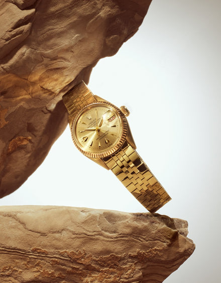 A gold wristwatch rests between two rocky surfaces, warmly lit against a plain background.