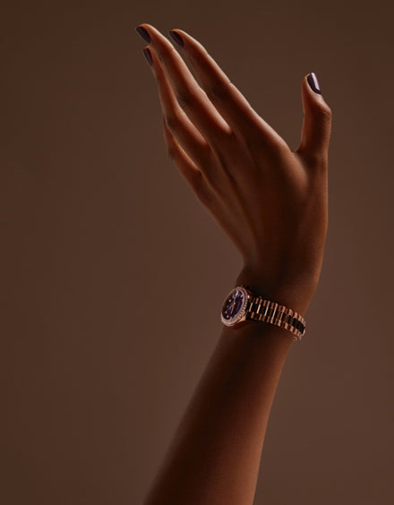 A raised hand with dark nail polish and a gold wristwatch against a brown background.