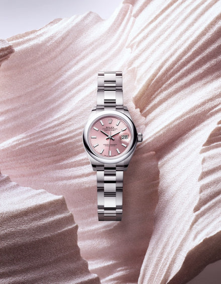 A silver Rolex wristwatch with a pink dial displayed on a textured, wavy, light-colored surface.
