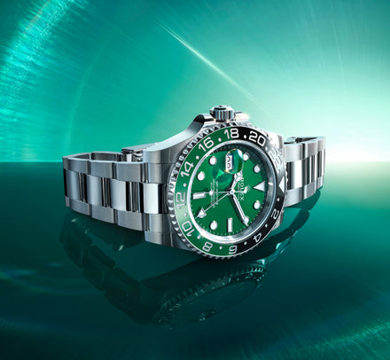 A luxury silver watch with a green face and black bezel displayed on a reflective green background.