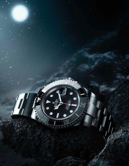 A silver wristwatch rests on rocks under a starry night sky with a bright full moon.
