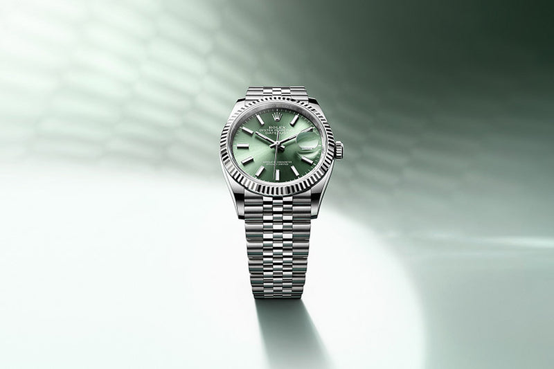 A silver wristwatch with a green dial and metal link bracelet on a light green, blurred background.