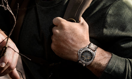 Close-up of a person wearing a wristwatch and holding a backpack strap, dressed in an olive green shirt.