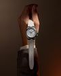 Wristwatch with white dial, cardinal point markers, dual time display, and light gray fabric strap worn on wrist.