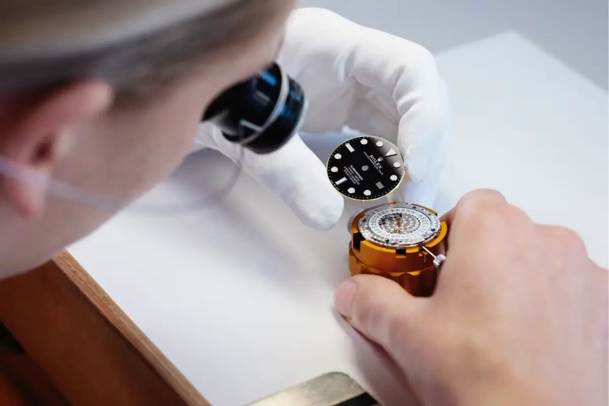 A watchmaker with gloves inspects and assembles the dial of a wristwatch at a workbench.