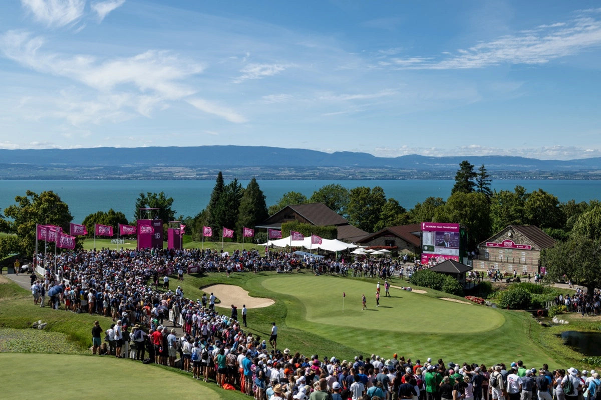 Large crowd watches golfers on a green near a lake, with mountains and blue sky in the background.