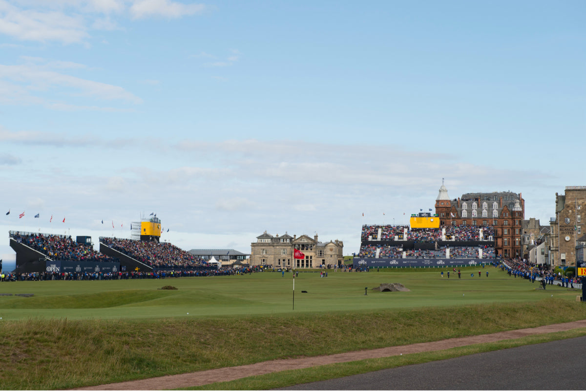 A large golf course with grandstands full of spectators and historic buildings in the background.