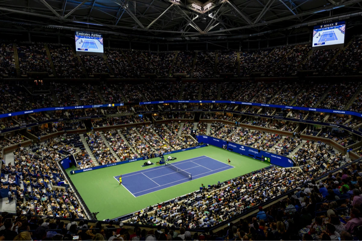 Crowded tennis stadium with a match in progress on a blue and green hard court under bright lights.