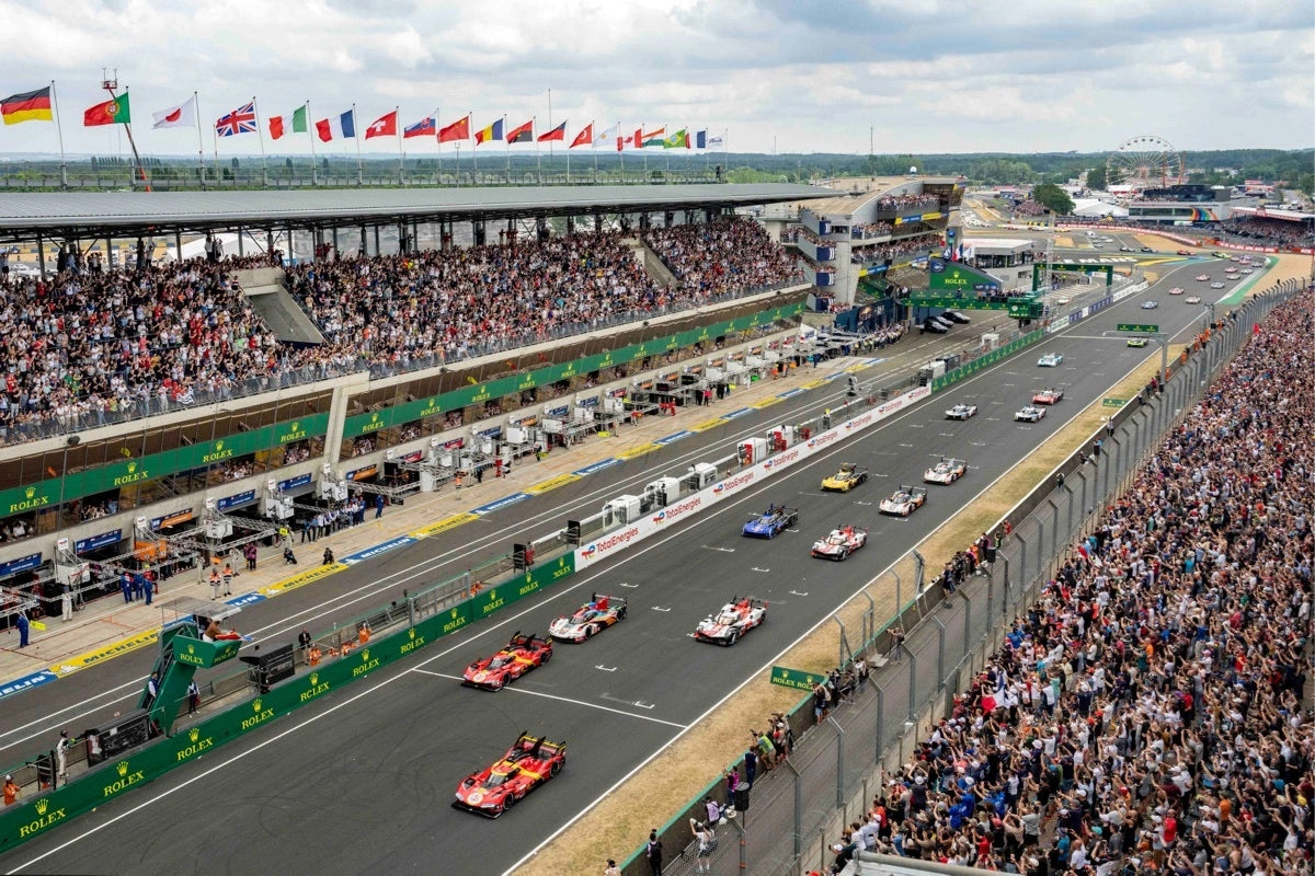 Race cars lined up on a track with grandstands full of spectators and flags overhead at a motorsport event.