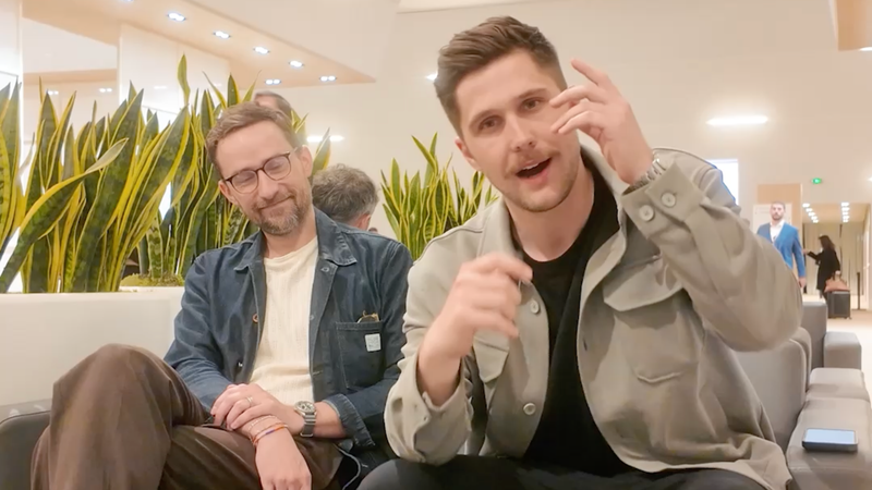 Two men sit on a couch indoors; one talks animatedly while the other smiles, with plants in the background.